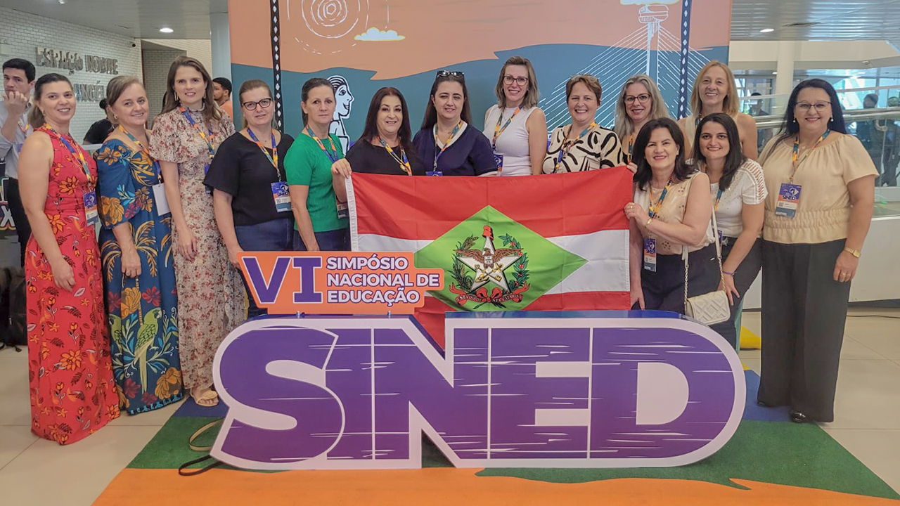 Foto de um grupo de 14 mulheres em pé. Duas seguram a bandeira de Santa Catarina. Na frente do grupo a logomarca do VI Sined em letras roxas.
