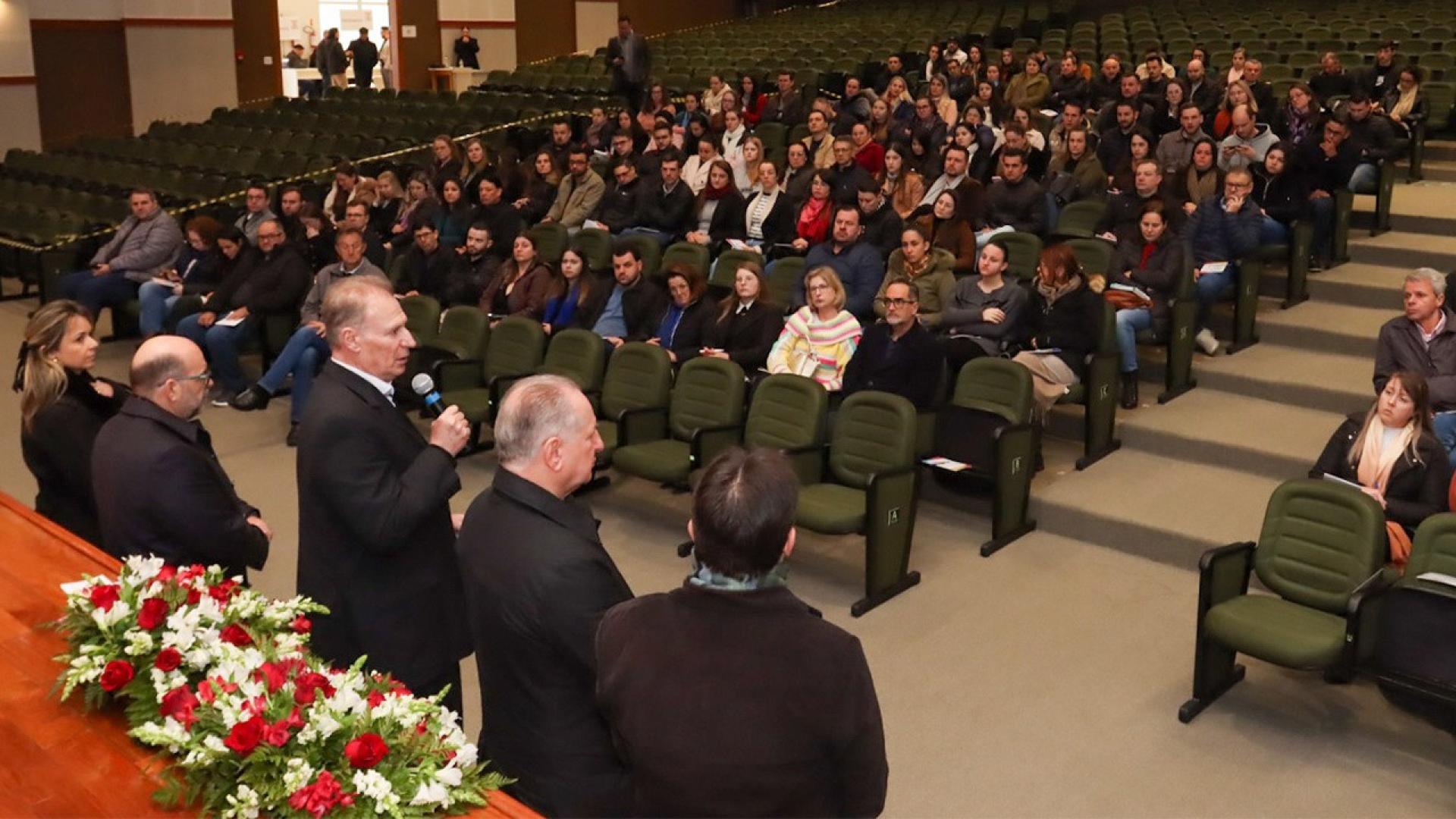 Foto do auditório. Na parte inferior, estão 4 homens e 1 mulher de frente para o público. O presidente do TCE/SC, conselheiro Herneus De Nadal, fala ao microfone.    