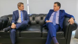 Foto do conselheiro José Nei Ascari e do deputado Rodrigo Minotto, sentados num sofá, conversando. Eles são homens brancos e vestem terno azul, com gravata também azul.