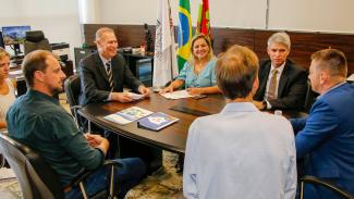 Foto com sete pessoas sentadas em volta de uma mesa, sendo duas mulheres e cinco homens. Na cabeceira esquerda, o presidente do TCE/SC. Ao fundo, as bandeiras do Brasil, de Santa Catarina e do Tribunal de Contas. 